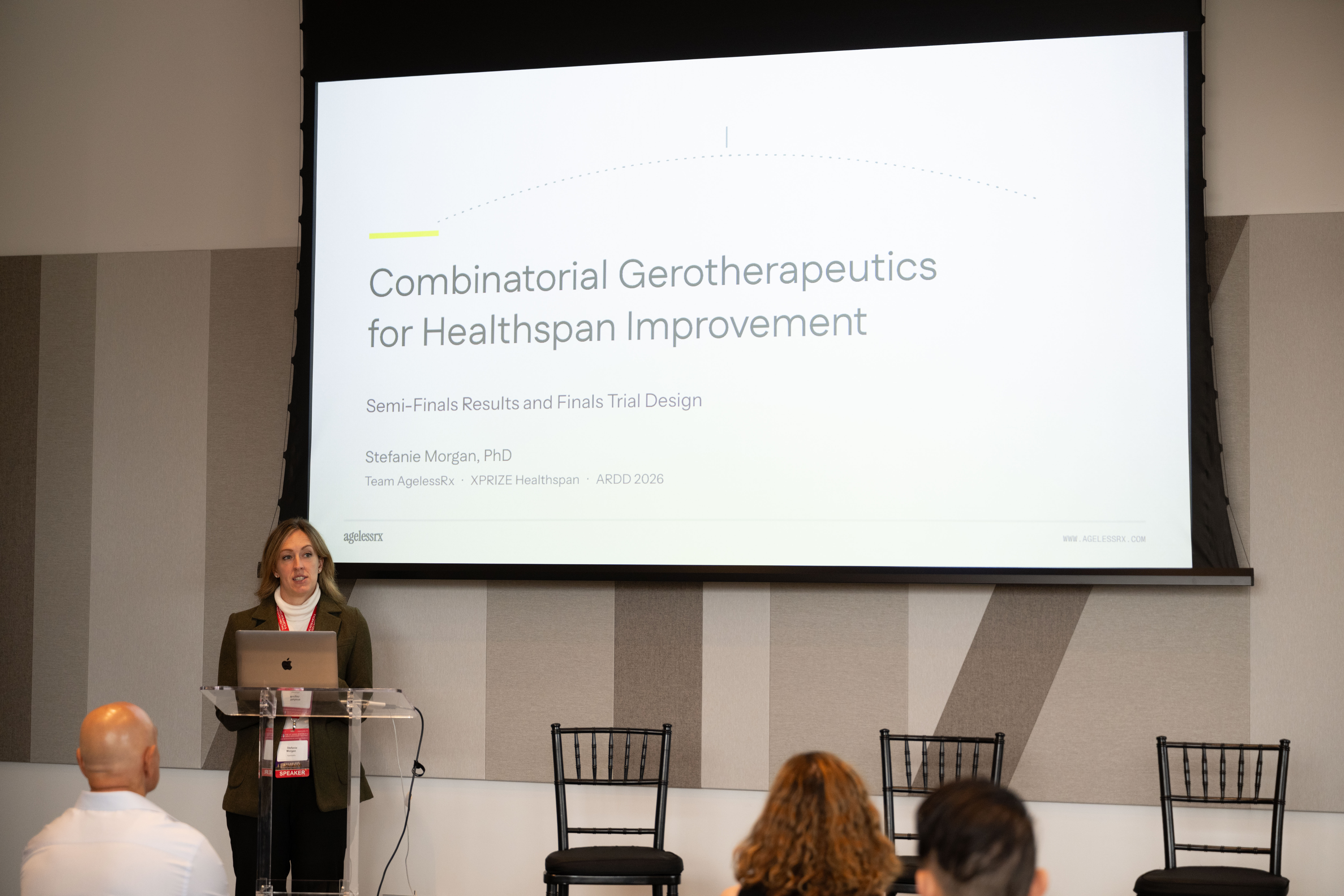 Stefanie Morgan at the podium beneath a title slide naming her and AgelessRx’s combinatorial gerotherapeutics presentation.