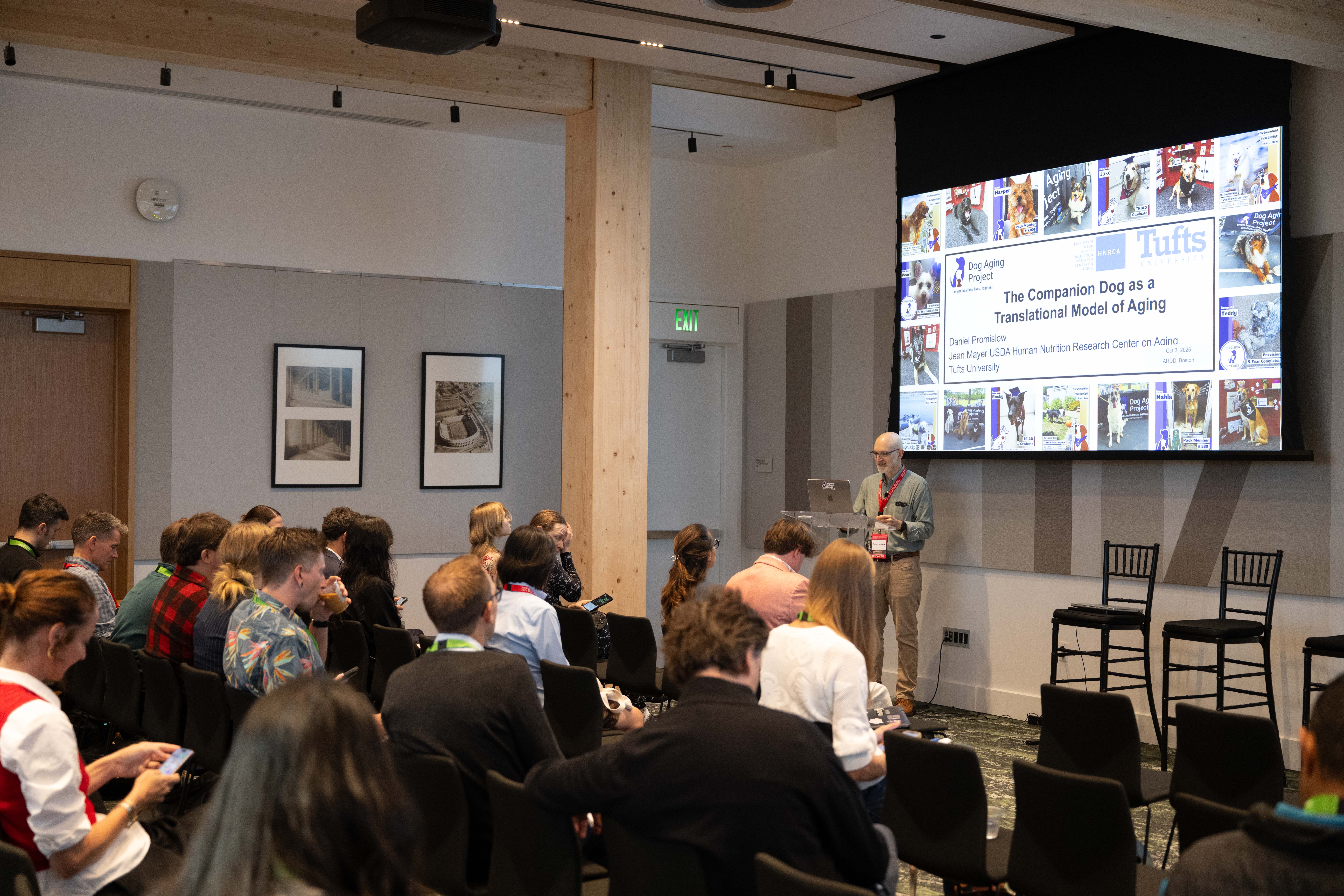 Daniel Promislow at the forum podium with a title slide naming him, Tufts University and The Companion Dog as a Translational Model of Aging.